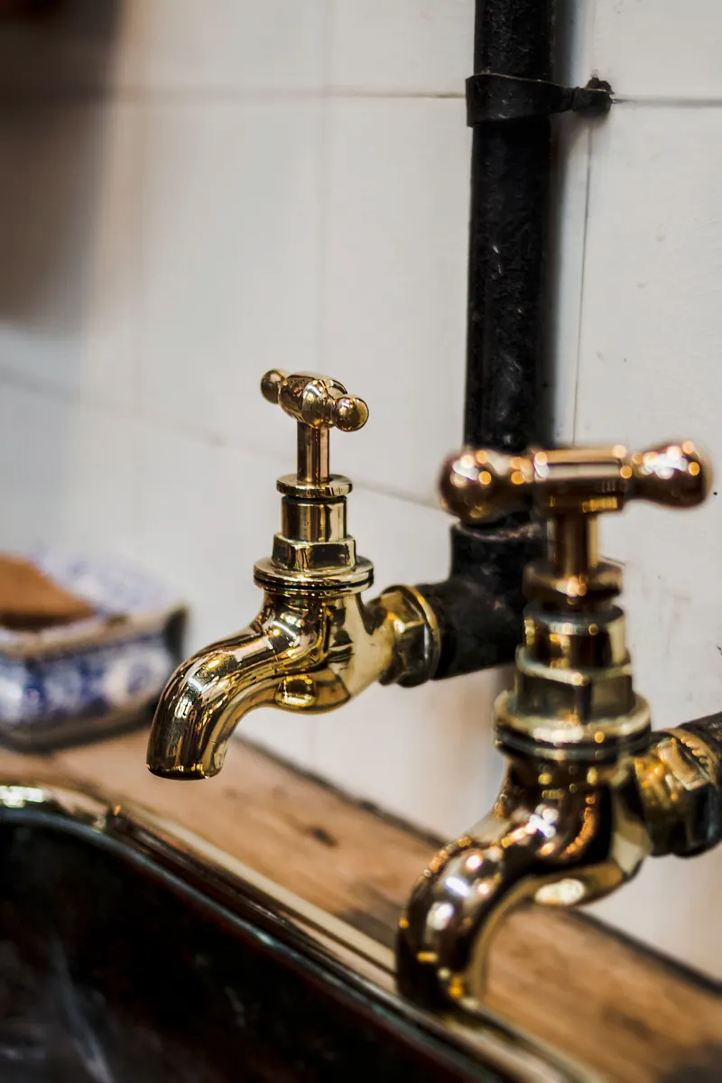 Duas torneiras de metal dourado sobre uma pia, ligadas a um cano preto na parede de azulejos brancos